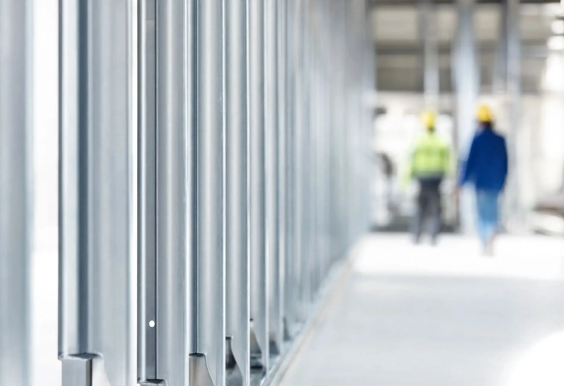 Rows of metal wall studs on an interior jobsite, with two tradespeople in hard hats walking in the distance